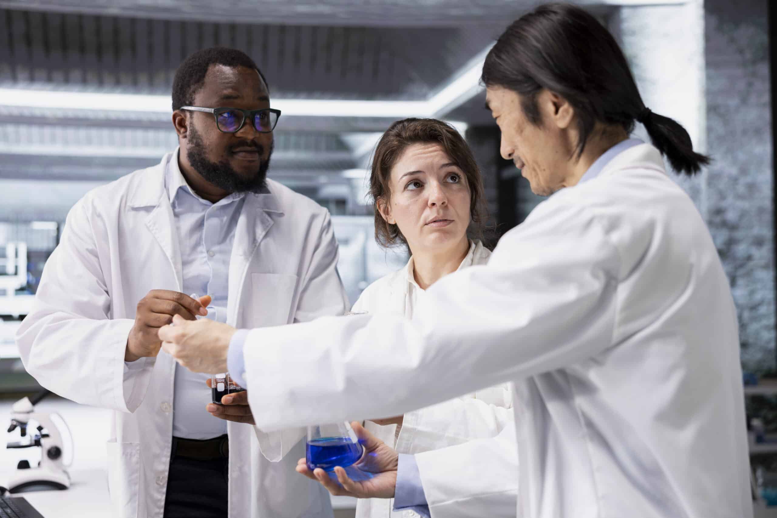 Team of lab chemists inspecting clarity of chemical solution in glass flasks for purity verification. Multiethnic research facility colleagues prepare reagent containers for assay procedures